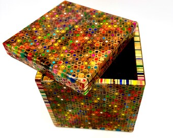 May include: A square box with a lid, crafted from the ends of colored pencils. The box's exterior is a mosaic of vibrant colors, with the pencil tips forming a textured surface. The lid is slightly ajar, revealing a black interior.