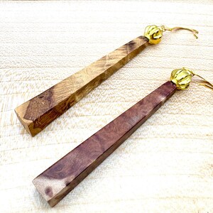 May include: A pair of long, wooden earrings with gold-toned metal accents. The earrings are made of different types of wood, one is a light brown wood and the other is a dark brown wood.