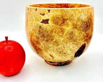 May include: A wooden bowl with a unique, swirling grain pattern. The bowl is light brown with darker brown swirls and has a smooth, rounded shape. A red apple sits next to the bowl.