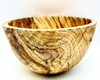 May include: A handcrafted wooden bowl with a natural, organic design. The bowl features a light brown color with darker brown and black grain patterns. The bowl's interior and exterior are smooth, showcasing the wood's natural texture. The bowl is ideal for serving food or as a decorative piece.
