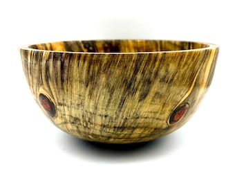 May include: A handcrafted wooden bowl with a natural, textured finish. The bowl features a light brown and dark brown grain pattern with two reddish-brown knots. The bowl is round and has a wide opening.