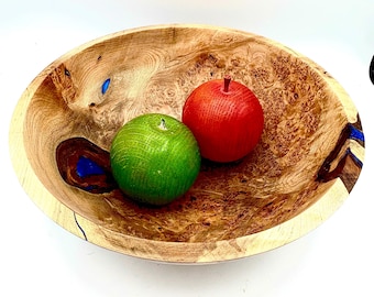 May include: A wooden bowl with a natural wood grain pattern, featuring a vibrant red and green apple. The bowl has blue accents and a smooth, polished finish. A decorative piece for the home.