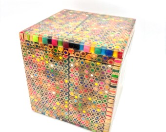 May include: A rectangular box crafted from the ends of colorful pencils. The box features a mosaic-like pattern of various colors, including red, yellow, green, and blue. The lid and sides are covered in the pencil ends, creating a unique and artistic design.