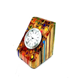 May include: A unique desk clock made from colorful pencils. The clock has a silver-colored face with black hands and Roman numerals. The clock's body is a rectangular prism shape, with the pencils arranged to create a mosaic effect. The clock measures approximately 4 inches in height.