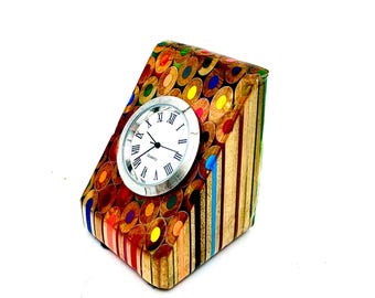 May include: A unique desk clock made from colorful pencils. The clock has a silver-colored face with black hands and Roman numerals. The clock's body is a rectangular prism shape, with the pencils arranged to create a mosaic effect. The clock measures approximately 4 inches in height.