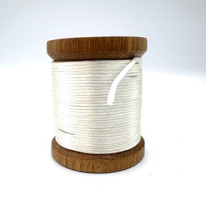 May include: A wooden spool with white thread. The spool has a dark brown, circular base and top, with the thread wound tightly around the center. A white thread end is visible.