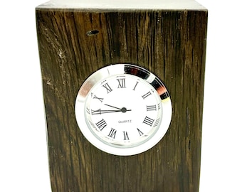 May include: A small, square wooden clock with a silver-colored bezel and a white clock face. The clock face features black Roman numerals and black hands. The wooden frame has a dark, textured finish.