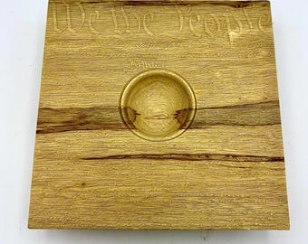 May include: A square wooden tray featuring a natural light brown wood grain. The surface is laser-engraved with the text We The People and Article I. A circular indentation is carved into the center of the tray, creating a smooth, concave bowl.