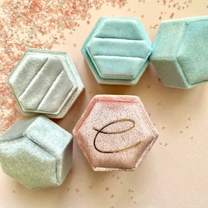 May include: A collection of hexagonal velvet ring boxes in shades of light blue and pink. One pink box features a gold letter "e". The boxes are arranged on a light pink surface, with scattered decorative elements.