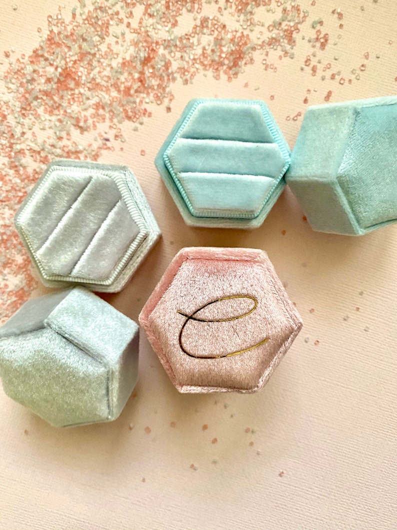 May include: A collection of hexagonal velvet ring boxes in shades of light blue and pink. One pink box features a gold letter "e". The boxes are arranged on a light pink surface, with scattered decorative elements.