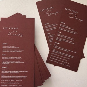 May include: Burgundy-colored menu cards with white text. The cards feature the words "Let's Feast" and individual names in elegant script. The menu items are listed below, including entrees, mains, and desserts.