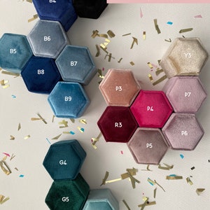 May include: A color chart of hexagonal velvet ring boxes in various shades of blue, green, pink, and beige. The boxes are labeled with color codes like B4, G4, and P3. The text "Box Colour Chart (cont'd)" is in the upper right corner. Gold confetti is scattered around the boxes.