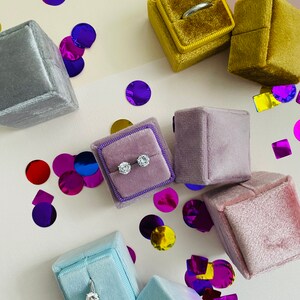 Könnte beinhalten: Sortiment von sechs Samtringboxen in verschiedenen Farben, darunter Lila, Grau, Blau und Gelb. Die Boxen sind mit Ringen gefüllt und von buntem Konfetti umgeben.