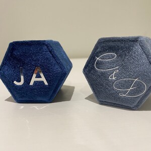 May include: Two hexagonal ring boxes in shades of blue. The box on the left has the initials "JA" in silver. The box on the right has the initials "C&D" in silver script. Both boxes are made of a soft, velvet-like material.