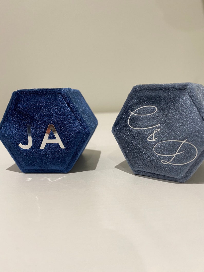 May include: Two hexagonal ring boxes in shades of blue. The box on the left has the initials "JA" in silver. The box on the right has the initials "C&D" in silver script. Both boxes are made of a soft, velvet-like material.