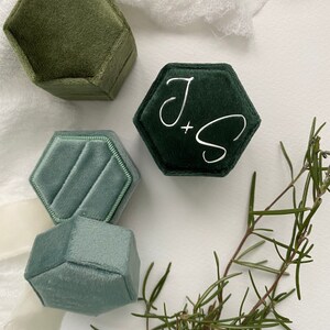 May include: Four hexagonal velvet ring boxes in shades of green and blue. One dark green box has the initials "J + S" in white script. A sprig of rosemary adds a natural touch to the arrangement. The boxes are empty.