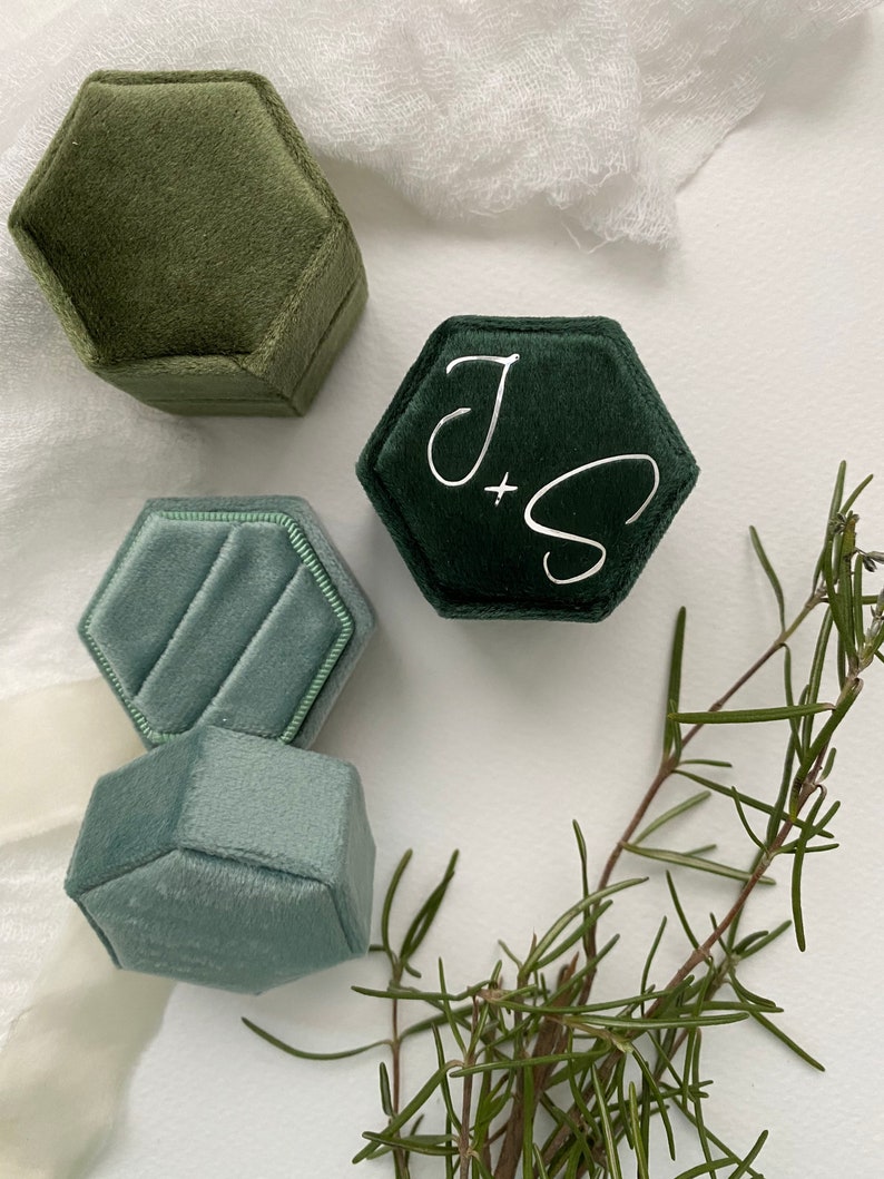 May include: Four hexagonal velvet ring boxes in shades of green and blue. One dark green box has the initials "J + S" in white script. A sprig of rosemary adds a natural touch to the arrangement. The boxes are empty.