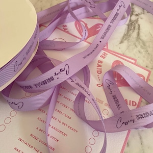 May include: Purple satin ribbon with the text "Cozys Bridal Shower" printed on it. The ribbon is wrapped around a white spool. The text "He Said..." is printed on a pink and white card.