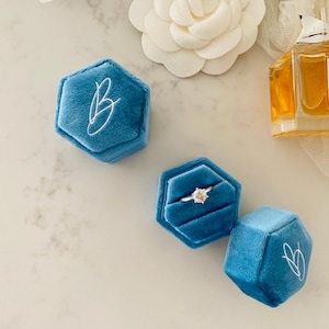 May include: Three hexagonal, teal velvet ring boxes with white script initials. One box holds a diamond engagement ring. A bottle of perfume and a white flower are in the background.