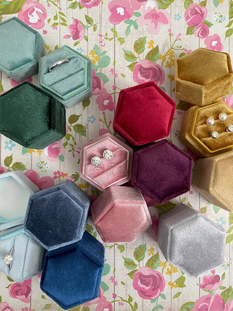 May include: A collection of hexagonal velvet ring boxes in various colors, including teal, green, red, gold, pink, and navy. Some boxes contain rings and earrings. The boxes are arranged on a floral patterned background.