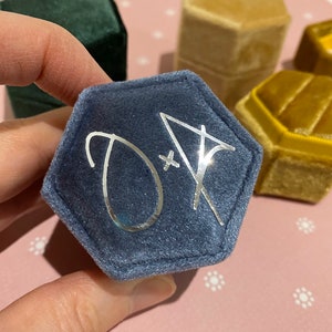 May include: A hexagonal, navy blue velvet ring box with silver initials "D + A" on the lid. Other velvet ring boxes in green and gold are visible in the background. The box is held by a hand.