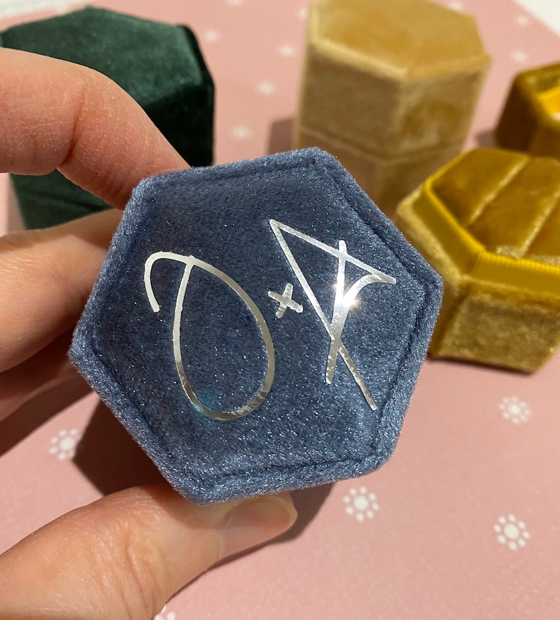 May include: A hexagonal, navy blue velvet ring box with silver initials "D + A" on the lid. Other velvet ring boxes in green and gold are visible in the background. The box is held by a hand.
