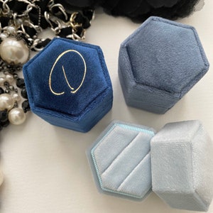 May include: A collection of hexagonal velvet ring boxes in shades of blue and gray. One box is navy blue with a gold letter. The boxes are open and closed, showcasing the interior. A black flower and pearl necklace are in the background.
