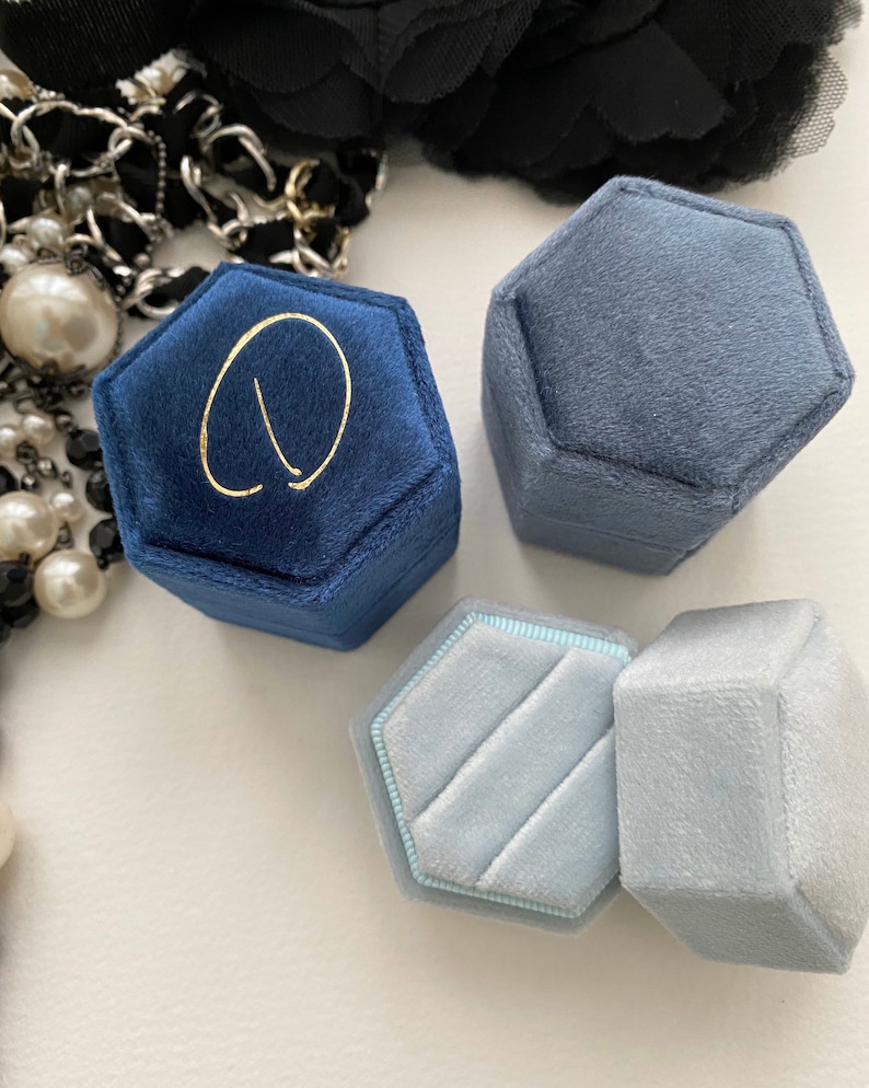 May include: A collection of hexagonal velvet ring boxes in shades of blue and gray. One box is navy blue with a gold letter. The boxes are open and closed, showcasing the interior. A black flower and pearl necklace are in the background.