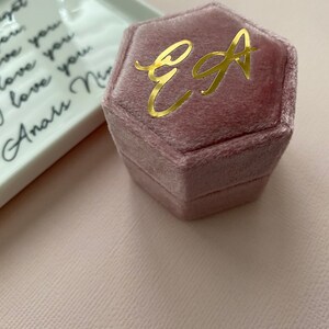 May include: A dusty rose velvet hexagonal ring box with gold lettering of the initials "EA". A white ceramic tray with black cursive text is in the background. The background is a light pink.