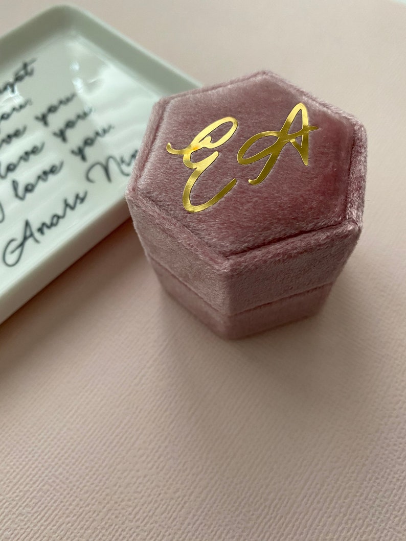 May include: A dusty rose velvet hexagonal ring box with gold lettering of the initials "EA". A white ceramic tray with black cursive text is in the background. The background is a light pink.