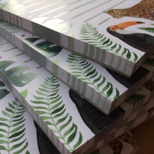 Op de afbeelding: Een stapel notitieboekjes met een tropisch thema. De covers bevatten aquarel illustraties van groene varens, monstera bladeren en een toekan. Het ontwerp bevat wit en groen gestreepte accenten en een luipaardprint patroon.