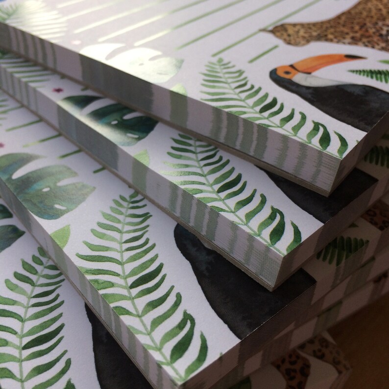 Op de afbeelding: Een stapel notitieboekjes met een tropisch thema. De covers bevatten aquarel illustraties van groene varens, monstera bladeren en een toekan. Het ontwerp bevat wit en groen gestreepte accenten en een luipaardprint patroon.