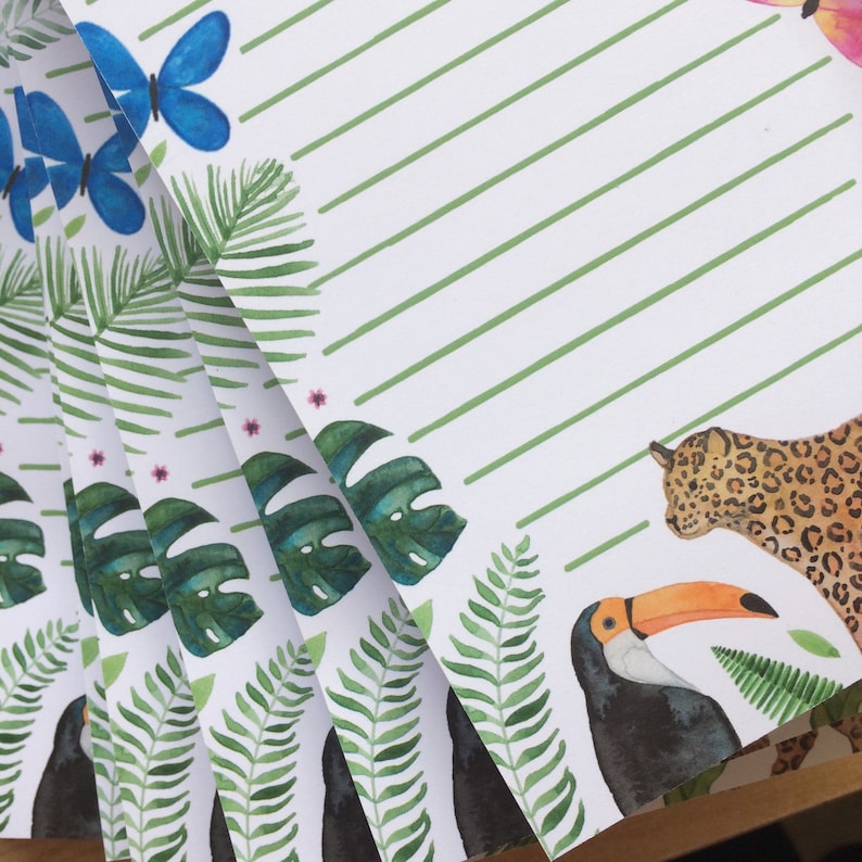 Op de afbeelding: Een stapel witte papieren vellen met aquarel illustraties van tropische planten, vlinders en dieren. De ontwerpen omvatten groene bladeren, een toekan, een luipaard en blauwe vlinders. Groene diagonale strepen zijn ook aanwezig.