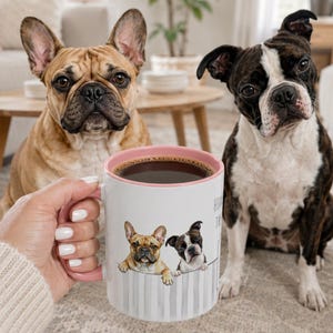 May include: A white ceramic mug with a pink interior and handle, featuring a printed illustration of a fawn French Bulldog and a black and white Boston Terrier peeking over a gray striped fence, filled with dark coffee.