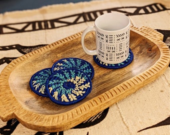 May include: Three blue and white woven coasters with a spiral design sit on a wooden tray with a white mug with a black geometric pattern.