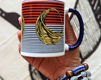 May include: White ceramic mug with a blue rim and handle. The mug features a colorful geometric design with red, yellow, and blue stripes and a yellow crescent shape.