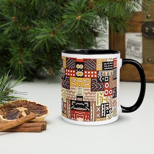 Puede incluir: Taza de cerámica blanca con borde y asa negros. La taza presenta un patrón geométrico colorido inspirado en el arte tribal africano.