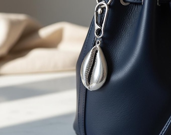May include: A dark navy blue leather bucket bag featuring a silver-toned metal cowrie shell charm attached to a lobster clasp. The bag has a drawstring closure with matching leather cords and silver-toned hardware accents on the side panel.