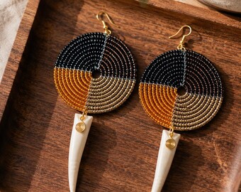 May include: A pair of circular earrings featuring concentric rows of black, gold, and bronze seed beads. Each earring has a gold-tone hook and a dangling, off-white, tapered horn-shaped pendant attached with a small gold spiral metal accent.