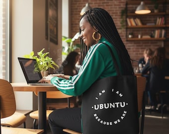 May include: A black tote bag with the text "I AM UBUNTU BECAUSE WE ARE" in a white circular design. The bag is carried by a person wearing a green jacket with white stripes, sitting at a wooden table with a laptop.