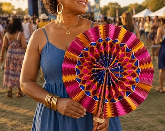 May include: A round, hand-held folding fan featuring a vibrant, geometric pattern in shades of magenta, orange, yellow, and blue. The fan is constructed with a pleated fabric surface and a wooden handle, showcasing a distinct, colorful design.