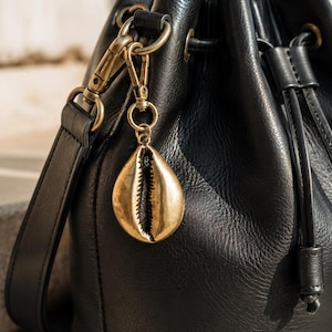 Könnte beinhalten: Eine schwarze Ledertasche in Beutelform mit Kordelzug und Schulterriemen. Ein metallischer, goldfarbener Kauri-Muschel-Anhänger ist mit einem messingfarbenen Karabinerverschluss und einem runden Ring an der Seite der Tasche befestigt.