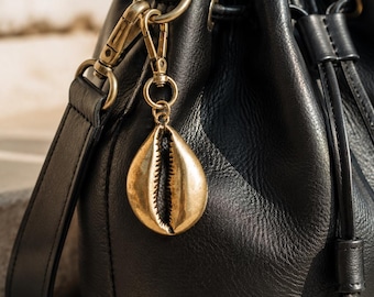 May include: A black leather bucket bag with a drawstring closure and a shoulder strap. A metallic gold-toned cowrie shell charm is attached to the side of the bag with a brass-colored lobster clasp and a circular ring, adding a decorative accent.