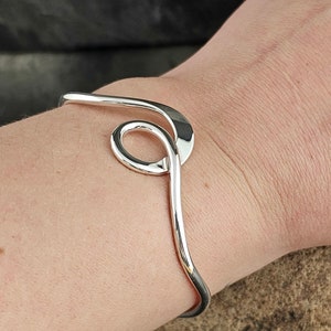 May include: A silver bangle bracelet with a unique, abstract design. The bracelet is made of a single piece of metal that is curved and twisted to create a flowing, organic shape.