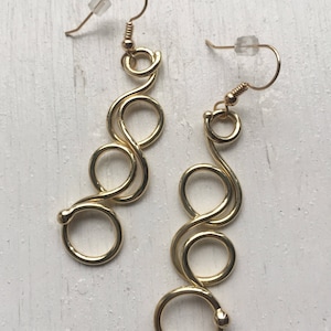 May include: A pair of gold-toned earrings with a unique, wavy design. Each earring features four interconnected circles, creating a flowing, abstract shape.