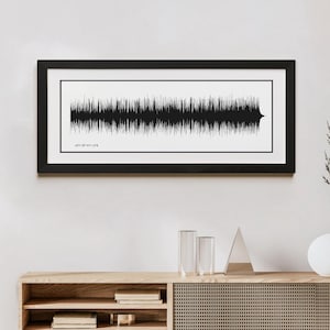 May include: A black framed print of a sound wave with the text "JOY OF MY LIFE" underneath. The print is hanging on a white wall above a wooden shelf with a vase and two glasses on it.