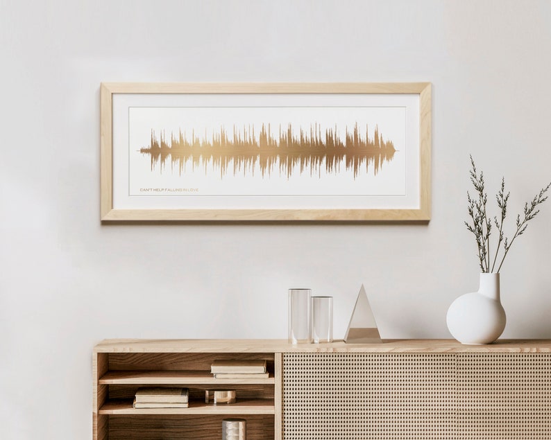 May include: A framed print of a gold sound wave with the text "Can't Help Falling In Love" underneath. The print is hanging on a white wall above a wooden cabinet with shelves.