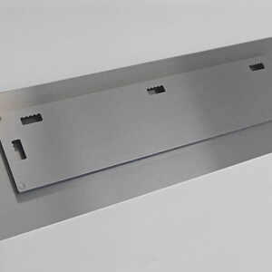 Op de afbeelding: De achterkant van een metalen wandkunstwerk met een ophangsysteem. Het stuk heeft een geborstelde zilveren afwerking en vier kleine gaten om op te hangen.