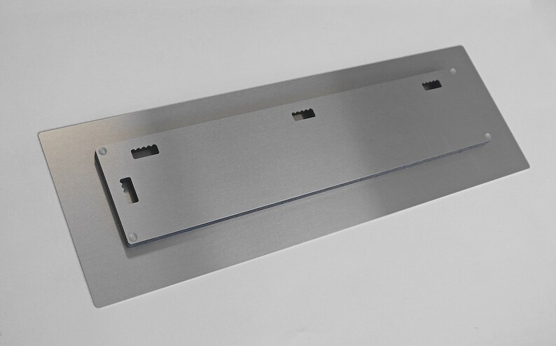 Op de afbeelding: De achterkant van een metalen wandkunstwerk met een ophangsysteem. Het stuk heeft een geborstelde zilveren afwerking en vier kleine gaten om op te hangen.
