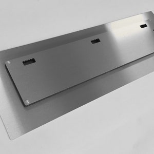 Op de afbeelding: De achterkant van een metalen wandkunstwerk met drie ophanggleuven. Het stuk is zilverkleurig en heeft een geborstelde afwerking.